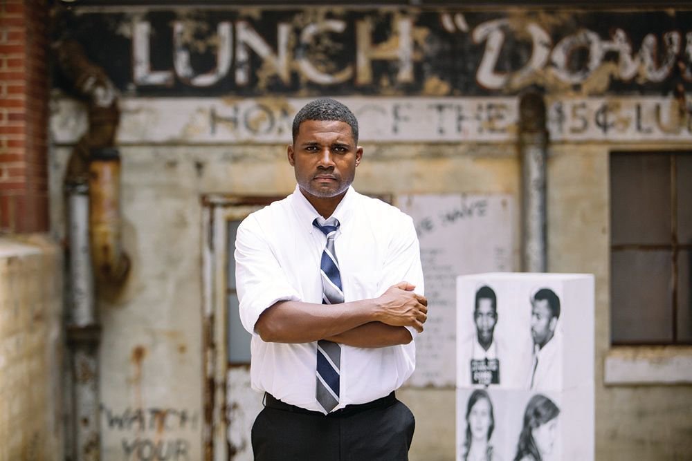 Photo of actor Mike Wiley, a black man wearing a white shirt, striped tie and dark pants. His arms are crossed in front of him.