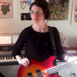 Woman in glasses holding an electric bass guitar. Keyboard behind her.