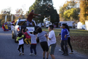 Photo of man standing in the bed of a truck making giant bubbles while several children follow behind