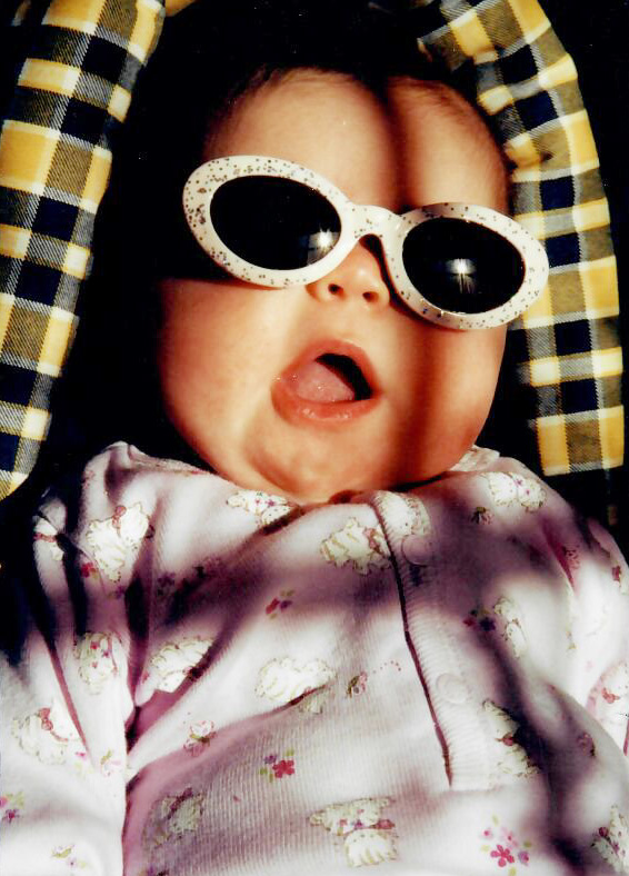 Photo of baby Lily McCoy Voller's face with sunglasses on and mouth open wide. The sun or stars are reflected in her sunglasses.