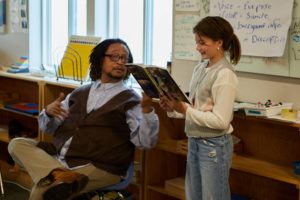 Photo of black man in sweater vest, seated, assisting a female student.