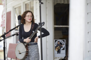 Photo of Sarah McCombie, standing on the Bynum Front Porch, holding a banjo, with one hand on her hip.