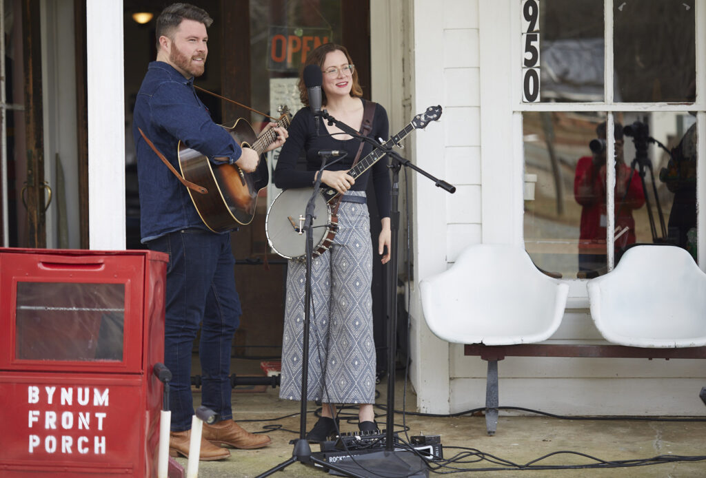 Photo of Austin and Sarah McCombie of the Chatham Rabbits, standing on Bynum Front Porch. Austin is holding a guitar and Sarah is holding a banjo. 