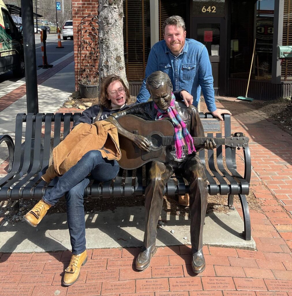 Photo of Sarah McCombie of the Chatham Rabbits sitting on a metal bench, posing with a statue of man playing a guitar. Austin McCombie is standing behind the bench, leaning on it. 