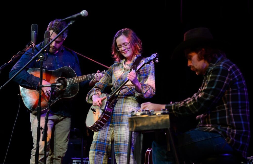 Photo of Chatham Rabbits playing live on a lighted stage. Sarah is playing the banjo, Austin is playing a guitar. A man is sitting at a steel guitar. 