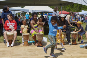 Photograph of a diverse group of people playing drums in a circle, with a small child dancing in the foreground