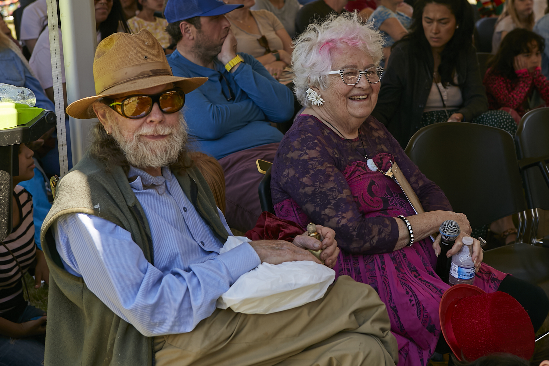 A bearded man wearing sunglasses and a hat (Snuffy Smith) and a woman with white hair and pink stripe, wearing a purple dress (Pam Smith) smiling at the camera