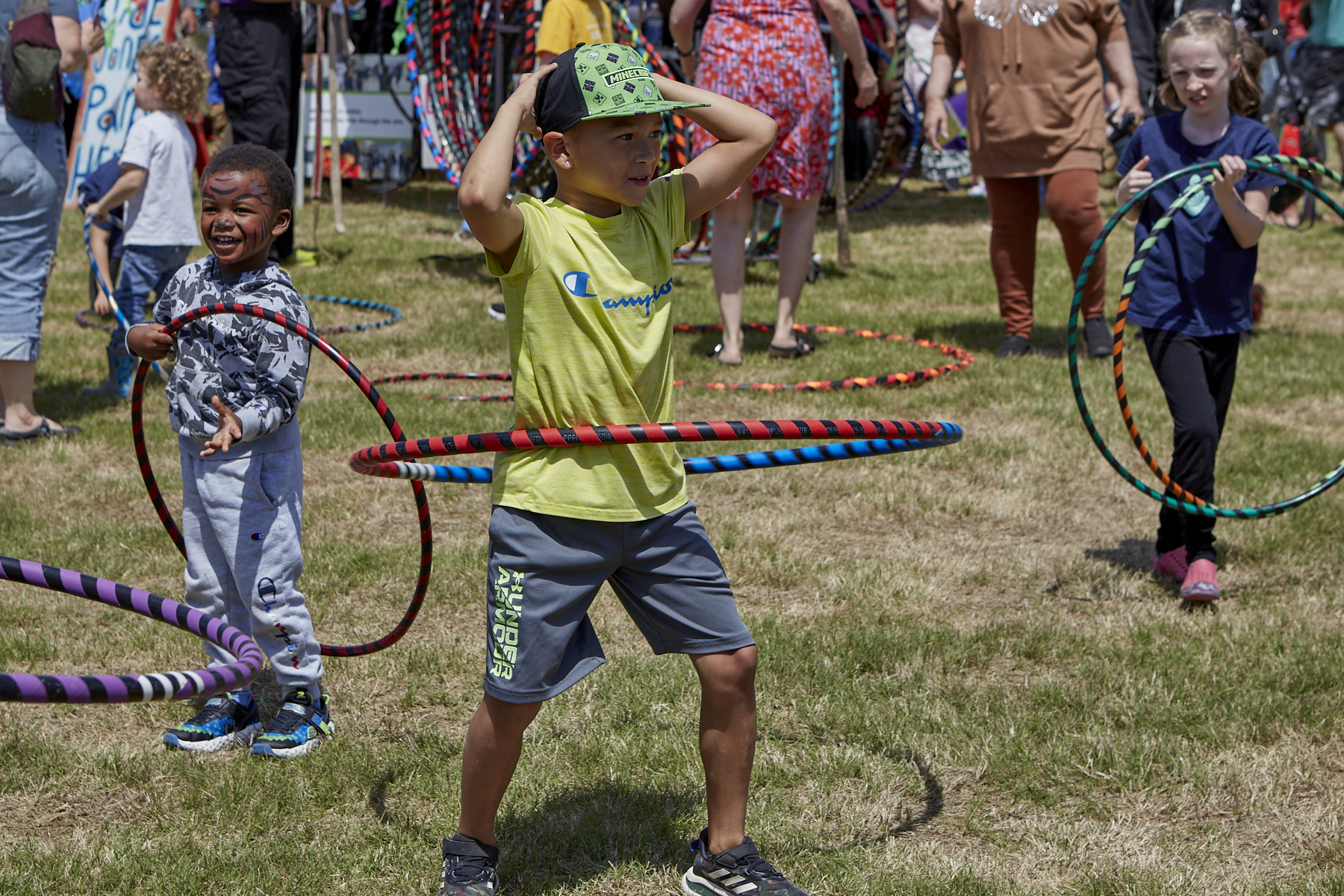 Photo from L to R: small boy with tiger face paint is holding a hoop; center - boy wearing a baseball cap is holding his arms above his head while hooping; R - a girl with pigtails carries 2 hula hoops