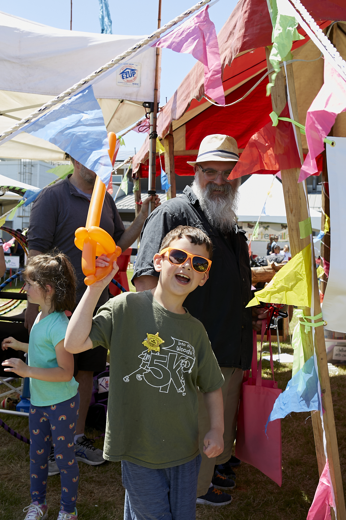 Photo of boy wearing sunglasses holding balloon sword in the air while bearded man (dad) smiles