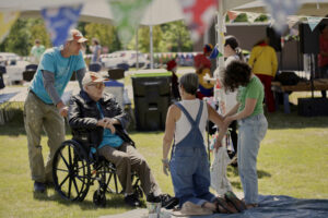 Photo: on left tall man pushing wheelchair with Clyde Jones; on the right a women in overalls kneels next to a young girl