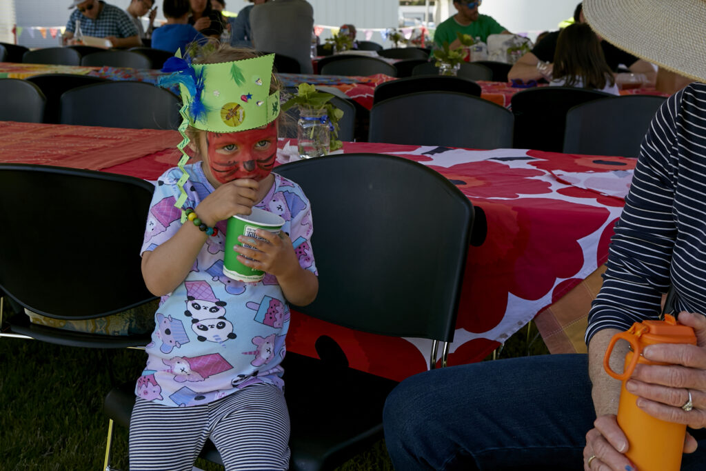 Photo of small child with a completely painted face and wearing a green paper crown eating ice cream