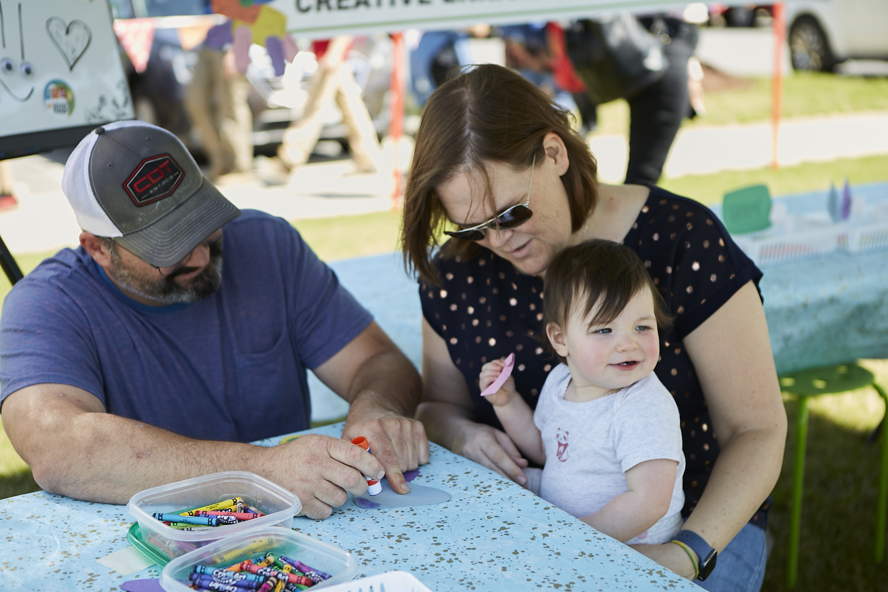 Photo of man in baseball cap holding a glue stick, a woman wearing sunglasses, holding a toddler and doing a craft