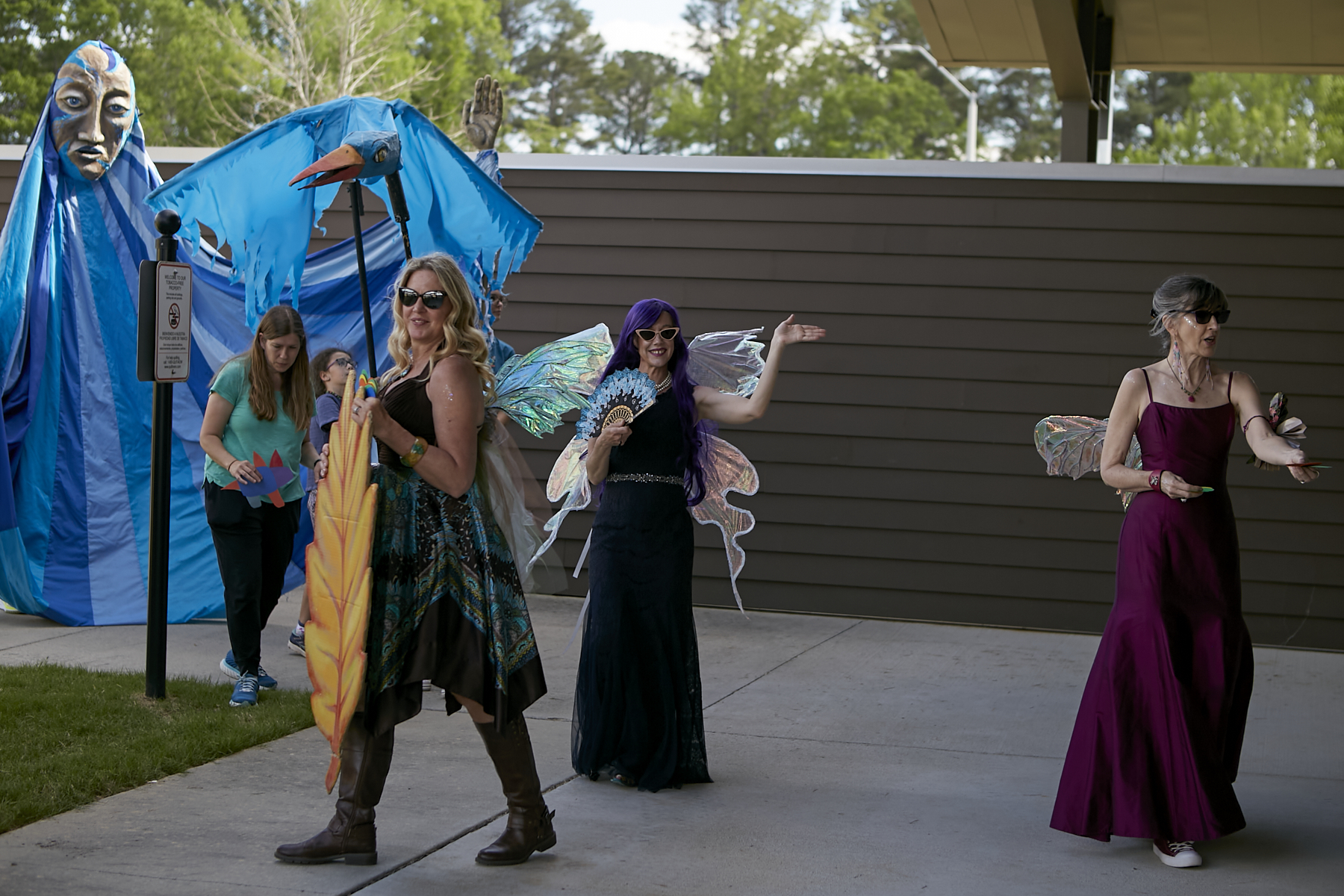 Photo of three women from WINGS community booth, all in beautiful gowns and wearing wings; left background giant blue puppet from Paperhand Puppet Intervention