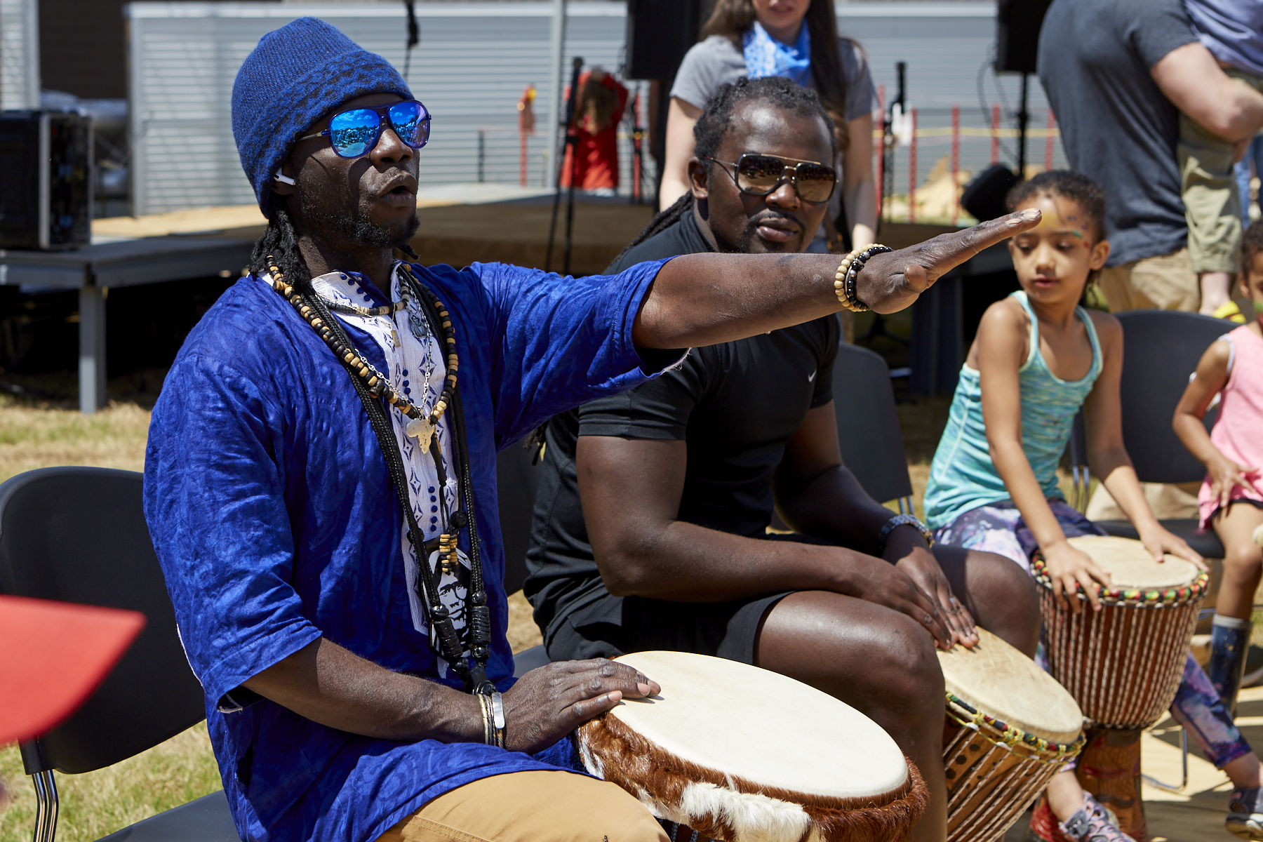 Photo of a man wearing a blue dashiki, sunglasses, and blue knit cap (Diali Cissokho) sits behind his drum and holds out his hand to demonstrate how to play, while a man wearing sunglasses, and a small girl, both with drums, watch