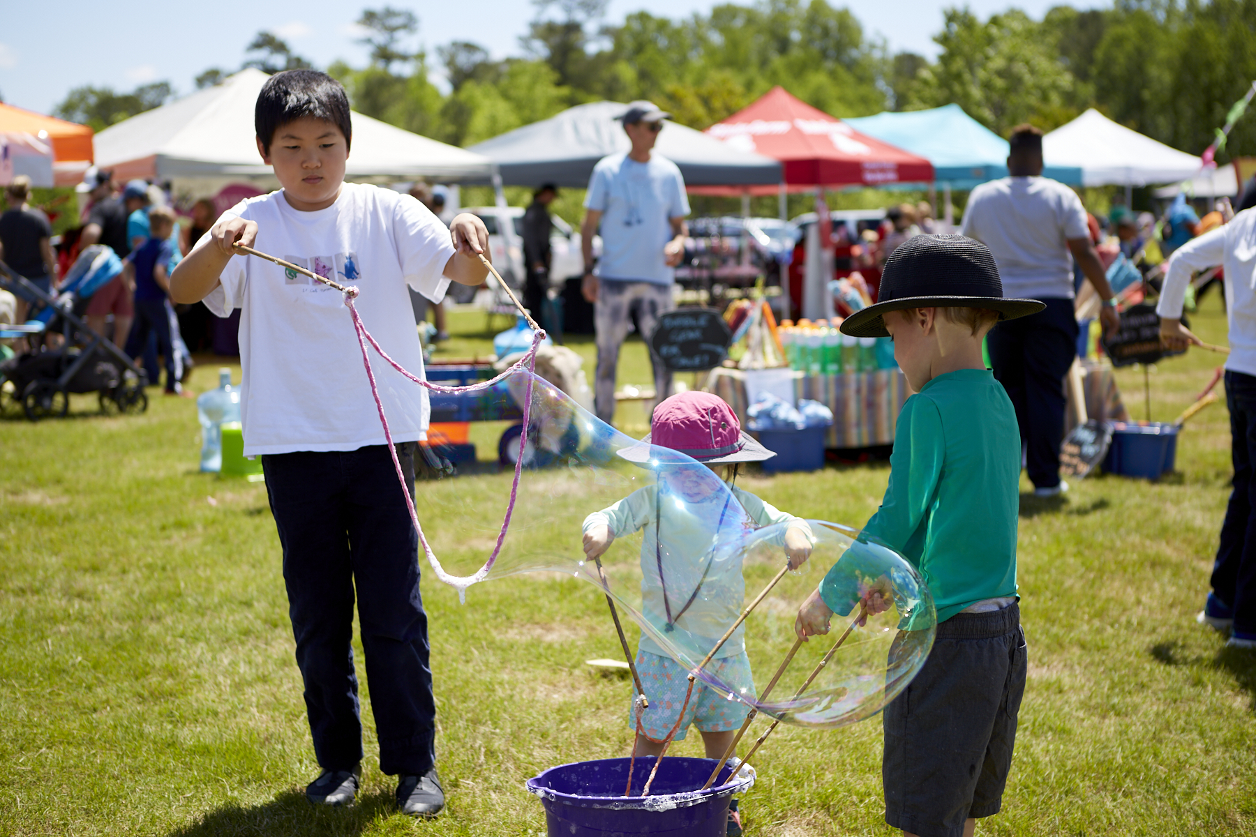 Photo of medium height boy, small girl in a sunhat, and a small boy in a sunhat making giant bubbles