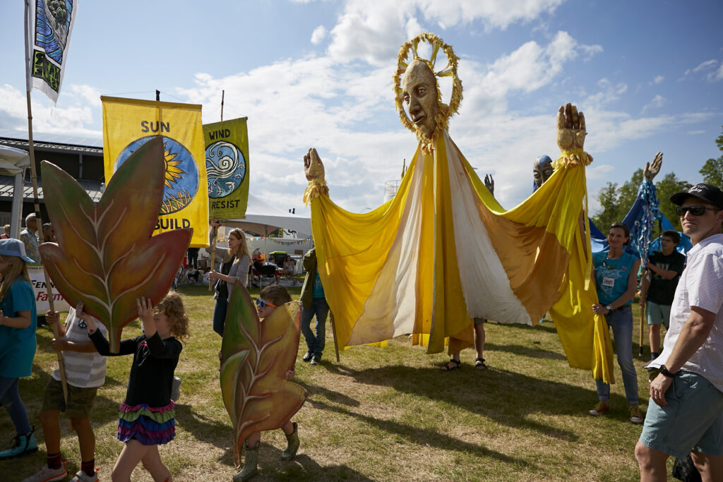 Photo of Paperhand Puppet Intervention's giant sun puppet and several other puppets and signs carried by festival-goers