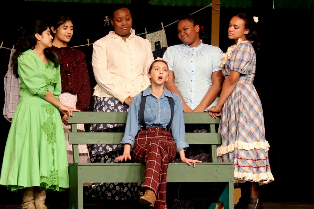 Photo of JMHS production of Oklahoma; 4 girls are standing behind a green bench where on girl is sitting and singing