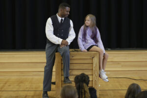 Photo of Mike Wiley and young girl sitting on a crate in performance of One Noble Journey