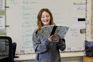 Photo of girl standing in front of a white board, holding a notebook and smiling