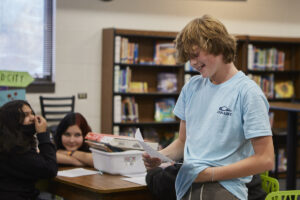 Photo of teen boy standing and reading his story while a seated girl in the background listens.