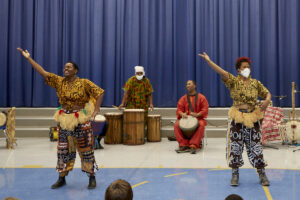 Photo - In front - 2 African women in traditional dress dance with their arms in the air; In Back two African men in traditional dress, one stands behind a trio of drums; one sits with a drum between his knees