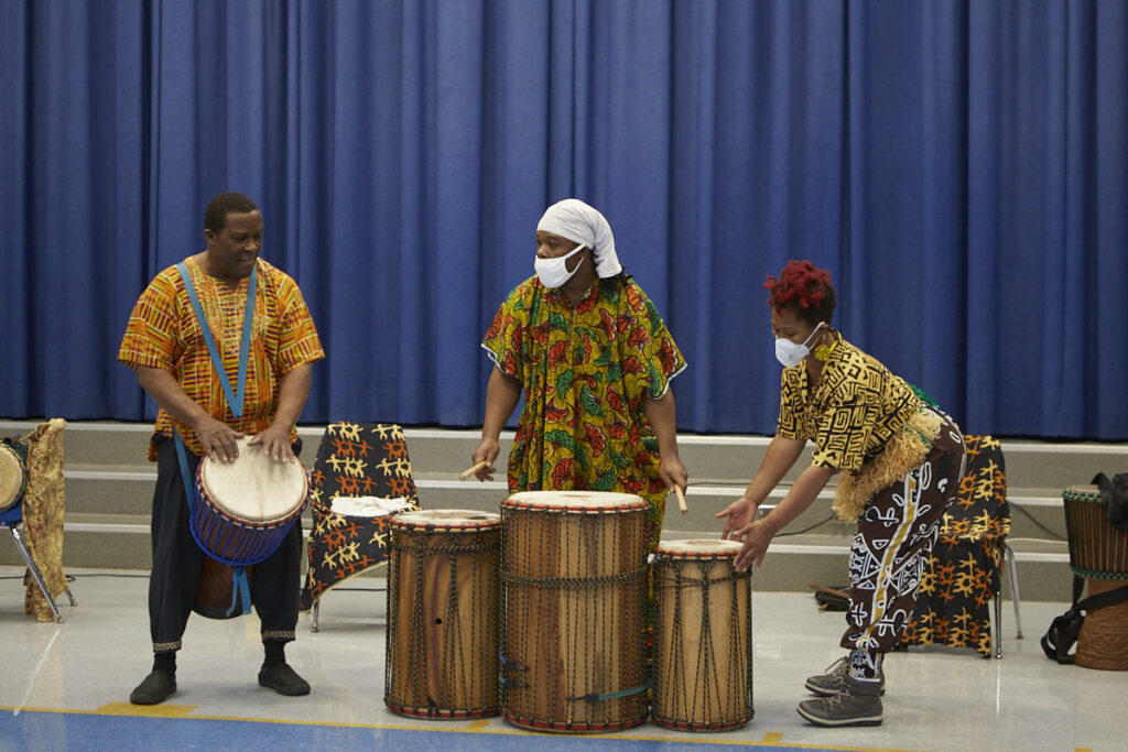 From L to R: African man in traditional dress playing an African drum; African man standing behind a trio of African drums; African woman in traditional dress playing drum