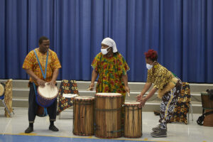 From L to R: African man in traditional dress playing an African drum; African man standing behind a trio of African drums; African woman in traditional dress playing drum