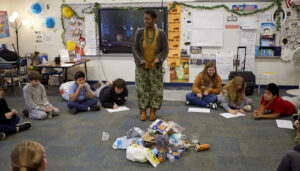 African woman standing in front of a pile of recycled items, with students sitting in a circle around her