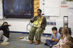 African woman sits holding a book pointing to a student, while students sit in circle around her