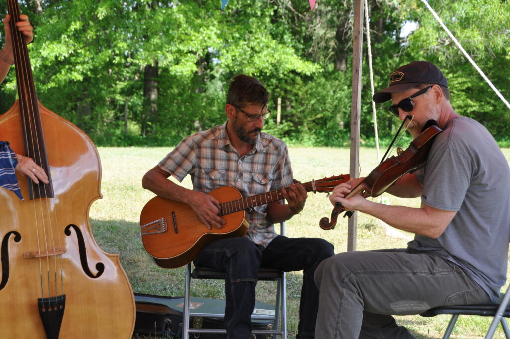 Three band members playing old-time music outside under a tent. Upright bass, guitar, and fiddle.