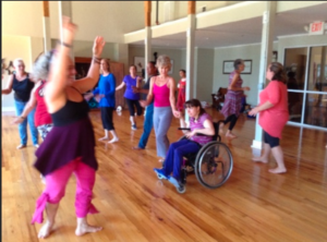10 to 15 women dancing in t-shirts and yoga pants in a large room. One woman with arms raised. One woman in a wheelchair.