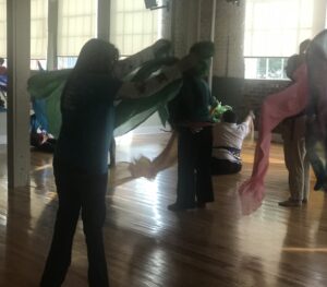 Five or six people in a large space with big windows, all dancing with silk scarves.