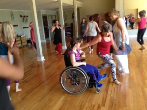 Nine or ten people in motion in tank tops and yoga pants. One woman moves quickly past another woman in a wheelchair.