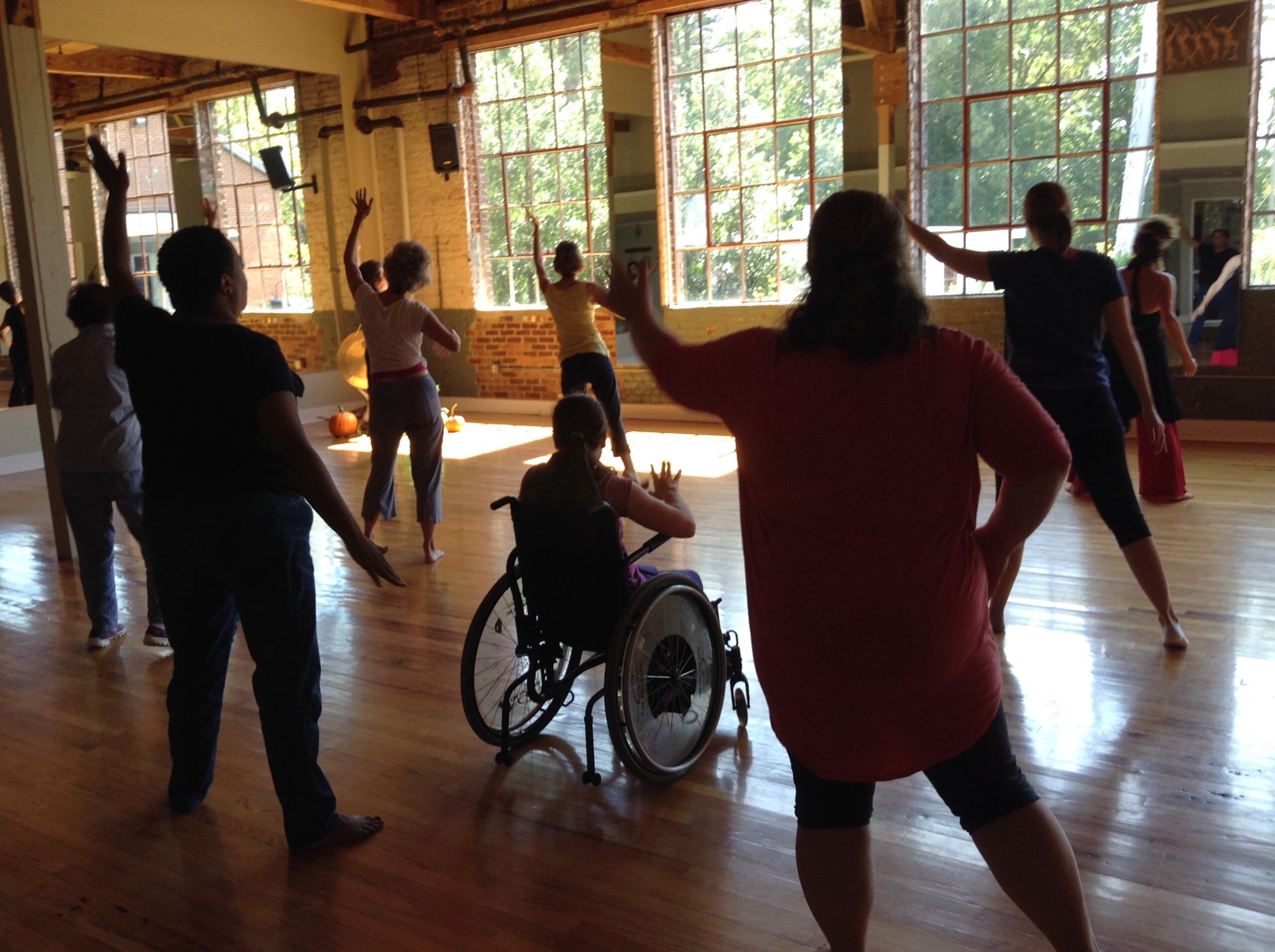 Nine people following choreography in a large converted mill dance studio. We see their backs. One participant in a wheelchair.