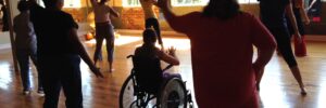 Nine people following choreography in a large converted mill dance studio. We see their backs. One participant in a wheelchair.