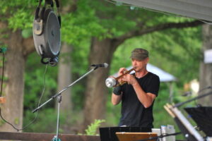 White man wearing a black t-shirt playing horn in an outdoor venue with trees in the background.