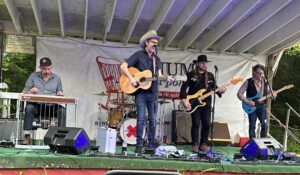 Five band members on an outdoor stage performing. Two wear cowboy hats.