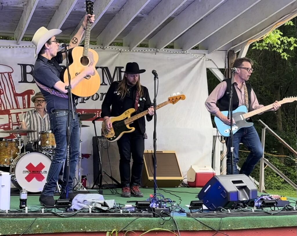 Four band members on an outdoor stage performing. Two wear cowboy hats.
