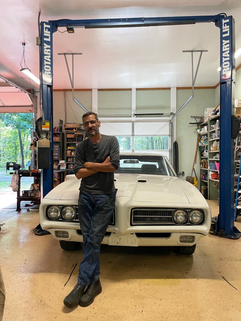White middle-aged man leaning against white Pontiac GTO in a metal shop.