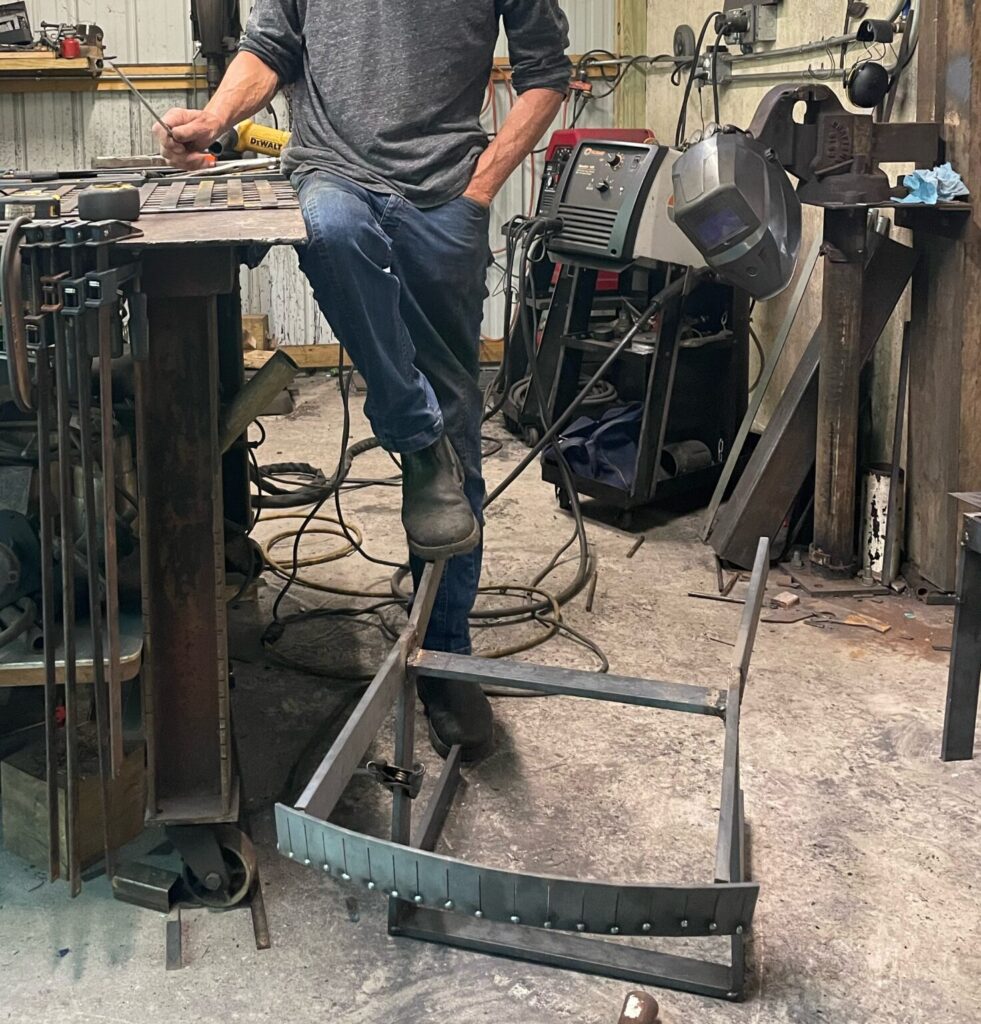White man in a gray shirt and jeans, leaning on his drafting table, with a foot resting on an in-progress stool creation.
