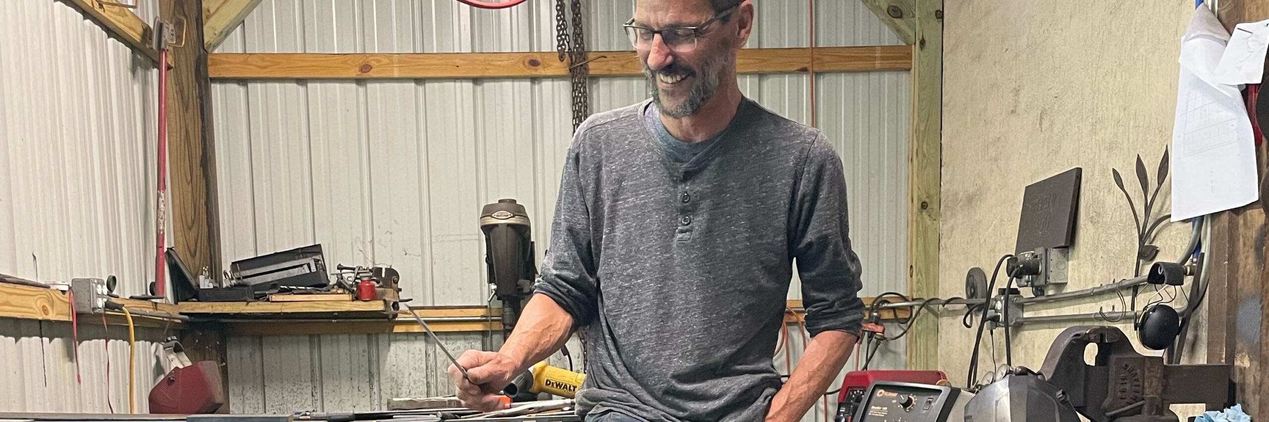 Smiling white man in gray henley shirt looking down at his drafting in his metal and woodworking shop.