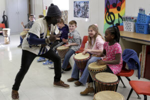 Diali stands in front of a girl encouraging her to play her drum