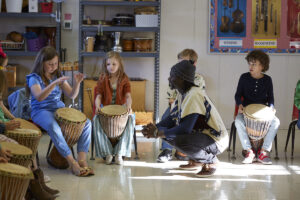Diali squatting next to a student, listening to her play her drum