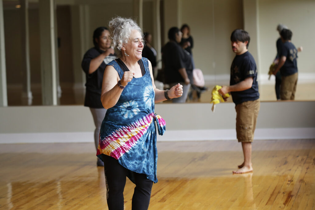 Photo of woman in tank top and yoga pants dancing with man and woman in background watching. 
