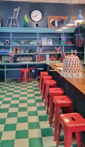Long counter lined with red metal stools. Green and tan checkered floor. Blue shelves holding small folk art pieces.
