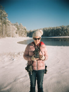 White womn with puffy jacket and winter hat standing beside a lake in the now. She is smiling and holding a camera.