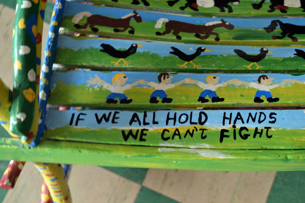 Close-up of wooden chair painted in folk art style. Green and blue with small figures of people, birds, horses. Painted text reads, "If we all hold hands . . ."