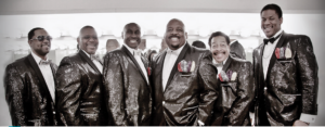 6 black men, members of Liquid Pleasure, stand smiling and wearing shiny tuxedos