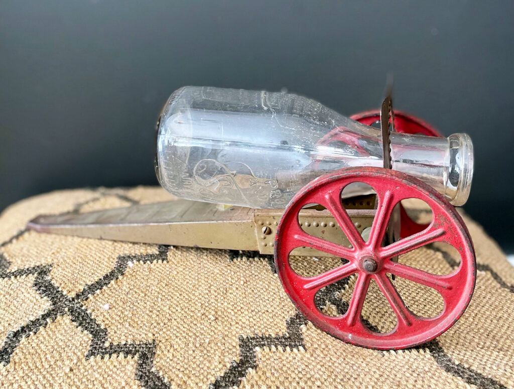 Sculpture: Small found metal ramp, two red metal spoke wheels, and a glass milk bottle.