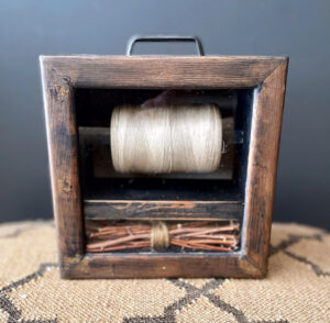 Sculpture: A small wood 3-D frame, with a spool of twine suspended within and small twigs in a bundle laying at the bottom.